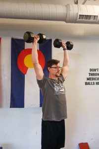 male personal trainer demonstrating a shoulder press with arms raised in the air